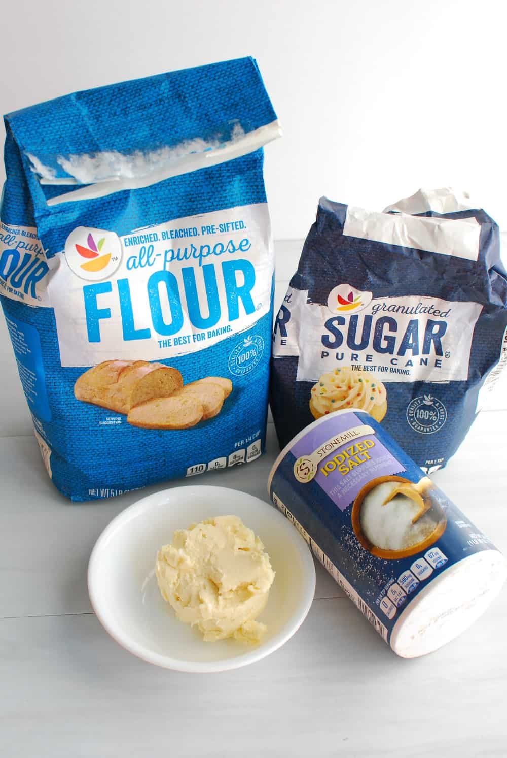 A bag of flour, a bowl of dairy free butter, a bag of sugar, and a canister of salt