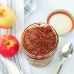 a jar of vegan apple butter next to a napkin, fresh apples, and a spoon