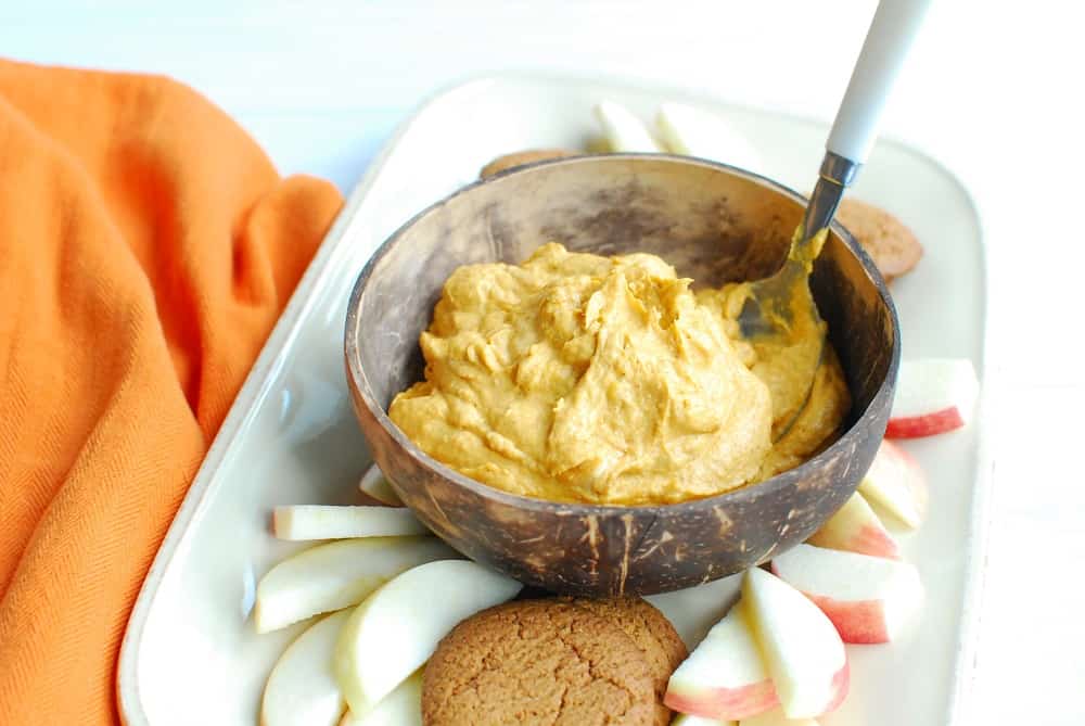 A bowl of dairy free pumpkin dip with a spoon in it, next to some sliced apples and gingersnap cookies.