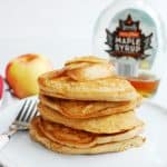 a stack of vegan apple pancakes on a white plate, next to a fresh apple and a bottle of maple syrup
