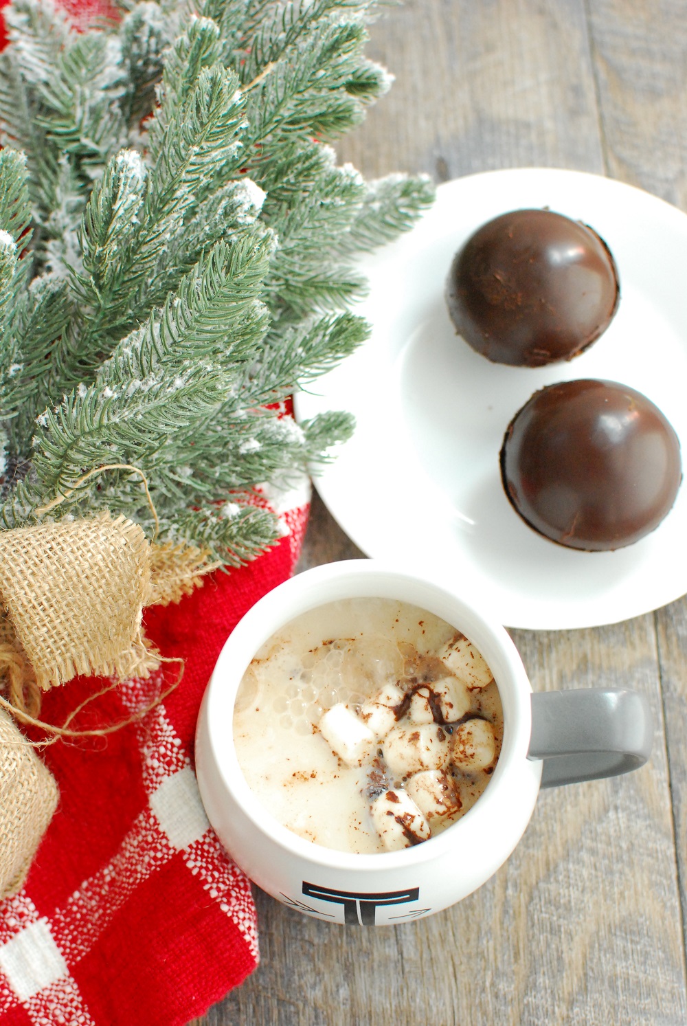 a mug of hot chocolate next to a few dairy free hot chocolate bombs and a fake mini christmas tree