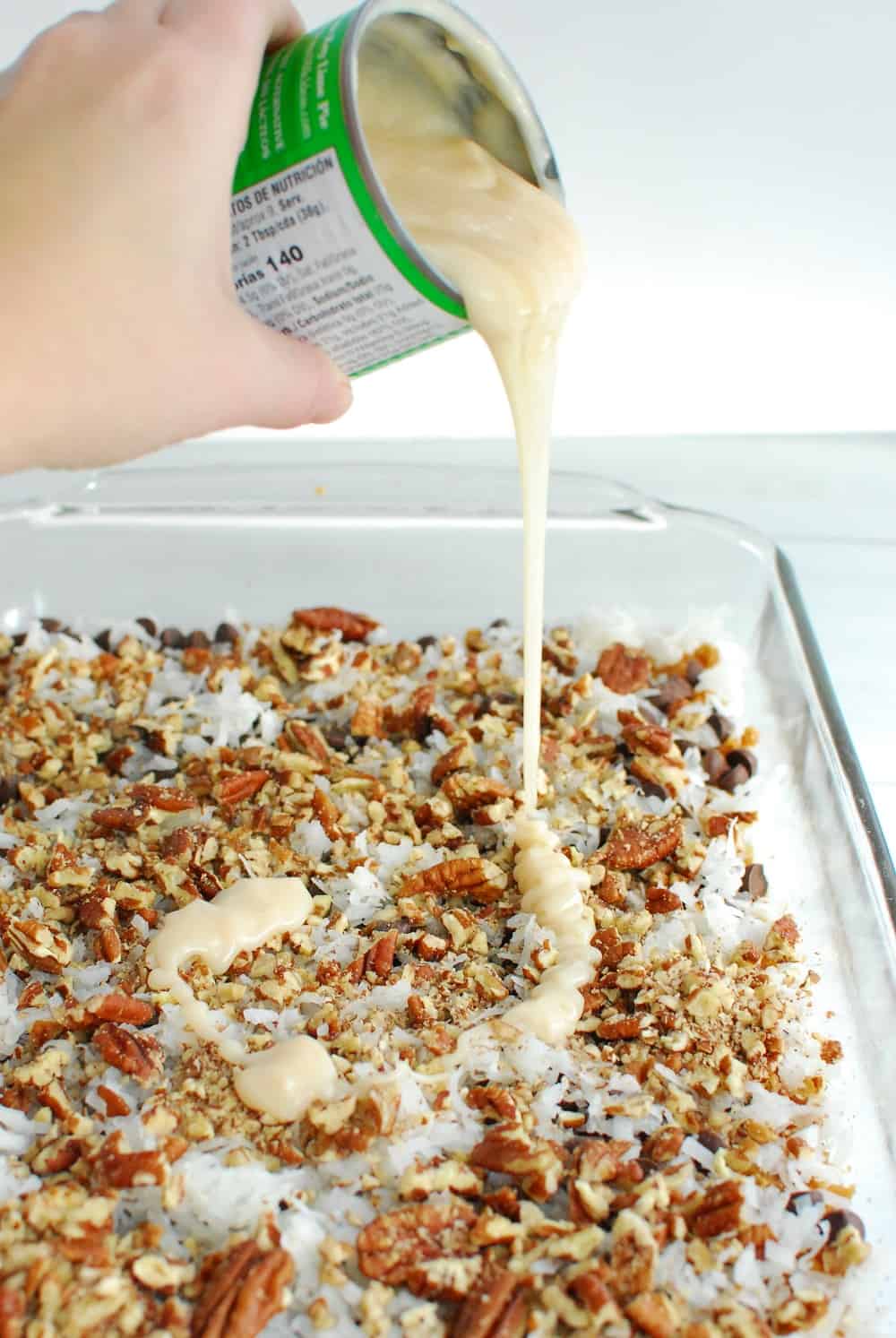 a woman pouring sweetened condensed milk into a baking dish with other ingredients