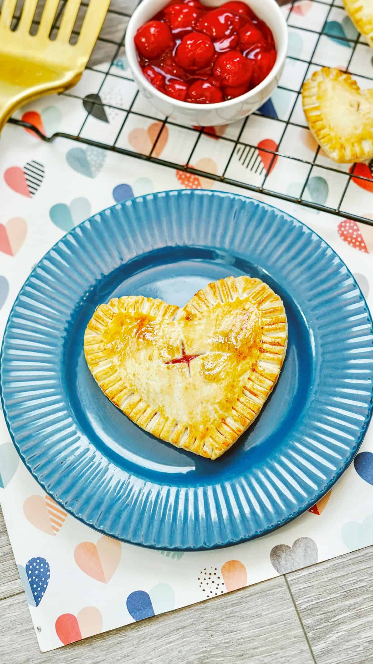 An air fryer hand pie on a blue plate next to a bowl of cherry pie filling and a spatula.