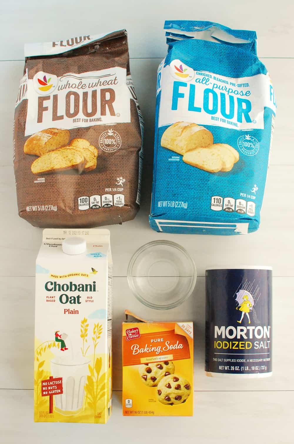 A package of whole wheat flour, a package of white flour, a carton of oat milk, a box of baking soda, a canister of salt, and a small bowl with vinegar.