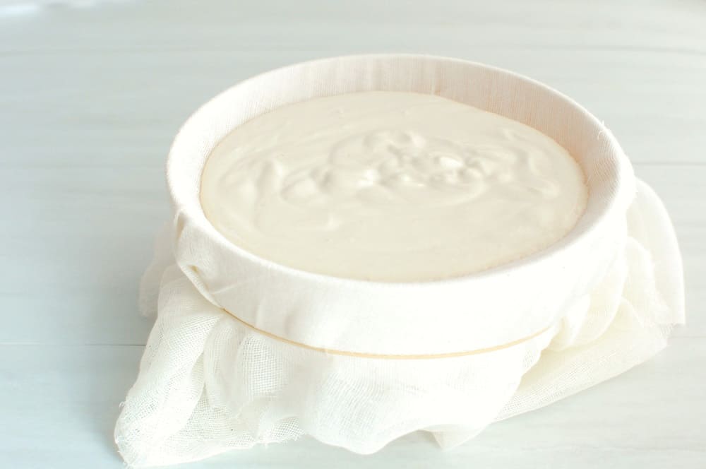 A bowl covered with cheesecloth, secured with a rubber band, with a yogurt mixture on top.