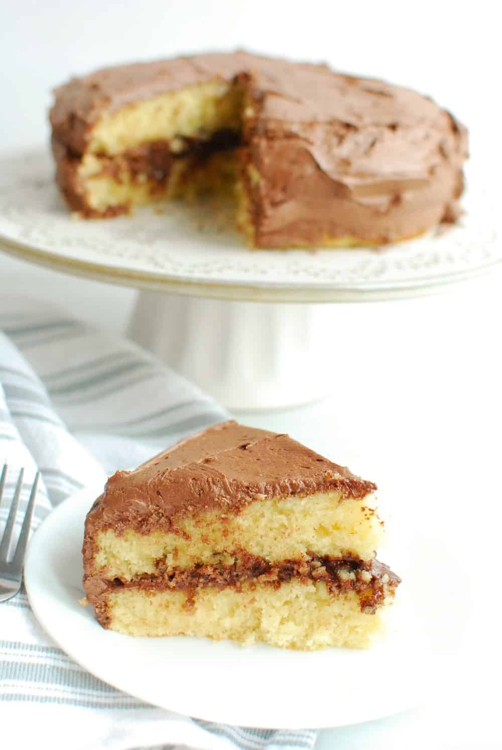 A slice of dairy free yellow cake with chocolate frosting on a white plate, next to a platter with the rest of the cake.