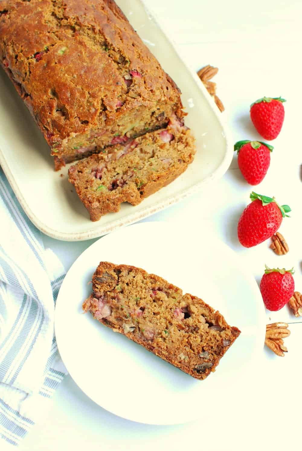 A slice of strawberry zucchini bread on a white plate next to the loaf and some fresh strawberries.