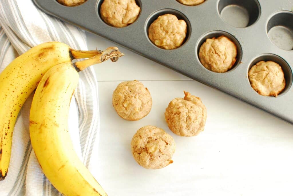 Some mini baby banana muffins on a table next to bananas and a napkin.