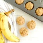 Three baby banana muffins next to some fresh bananas and a muffin tin.