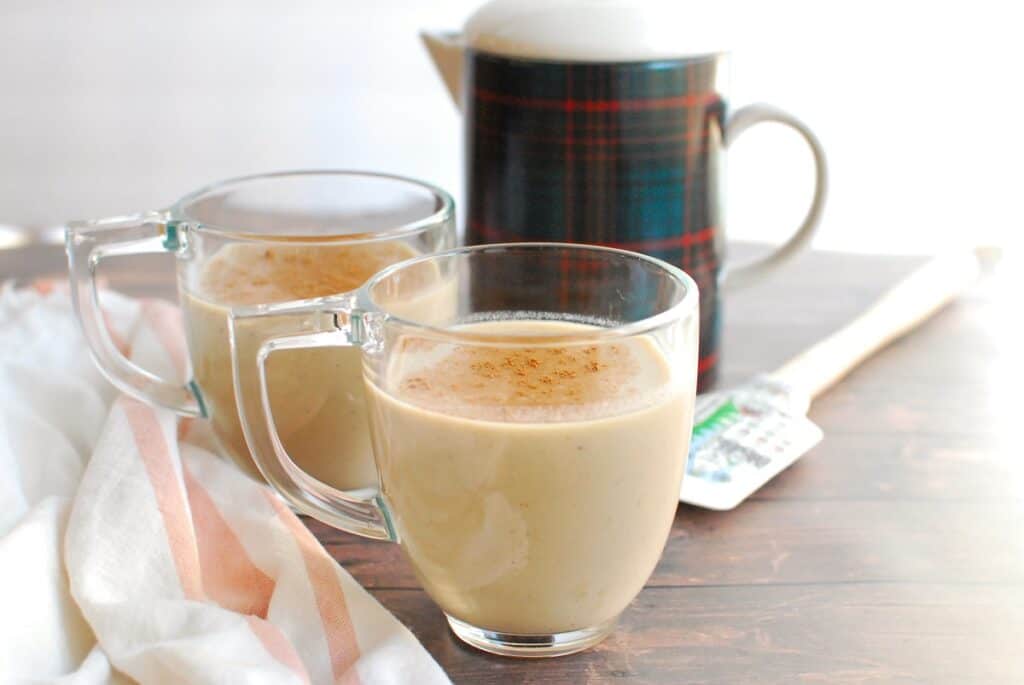Two mugs of dairy free eggnog next to a napkin and a pitcher.