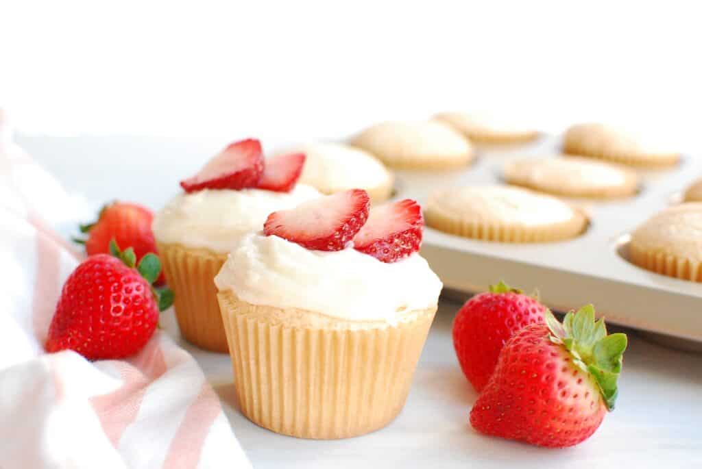 Two angel food cupcakes next to some fresh strawberries and a pan of cupcakes.