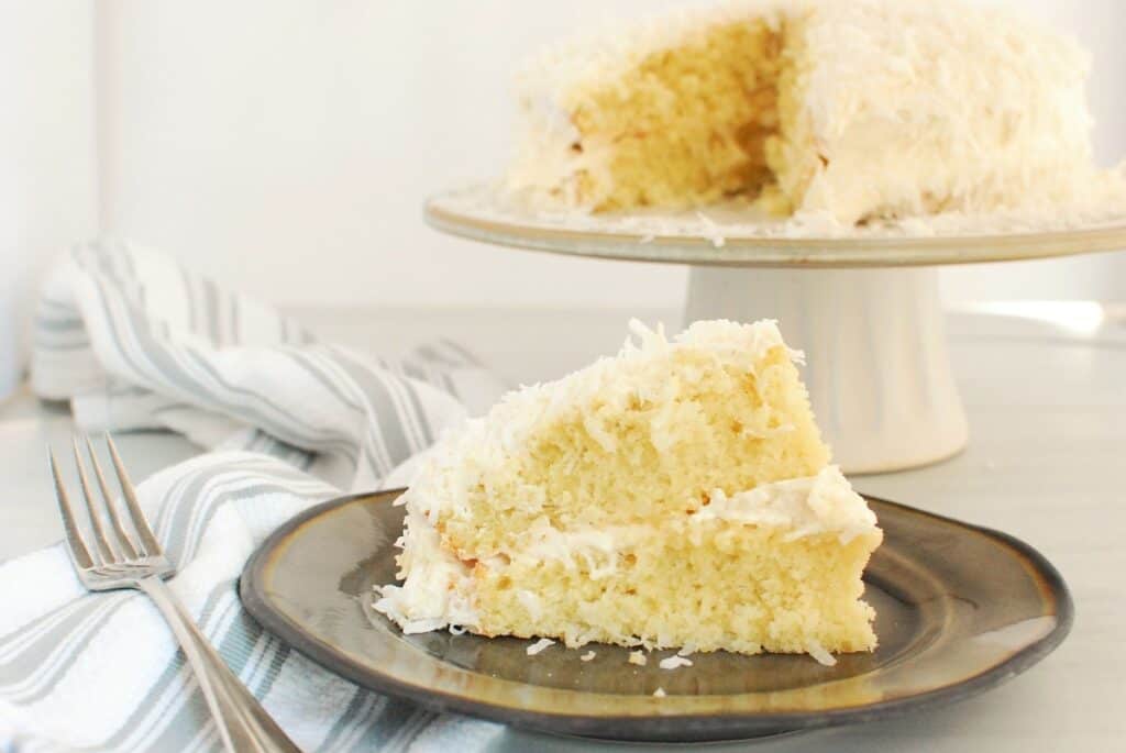 A slice of dairy free coconut cake on a plate with the full cake in the background.