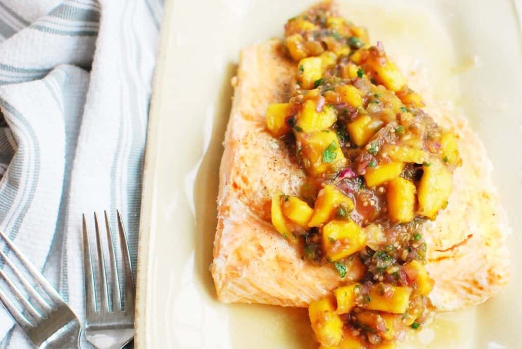 A piece of dairy free salmon topped with mango tomatillo salsa on a plate next to a fork.