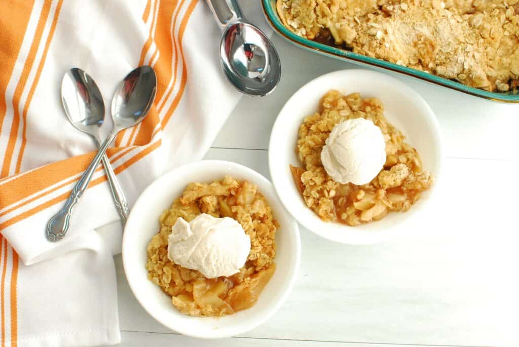 Two bowls of dairy free apple crisp next to spoons, a napkin, and a baking dish.