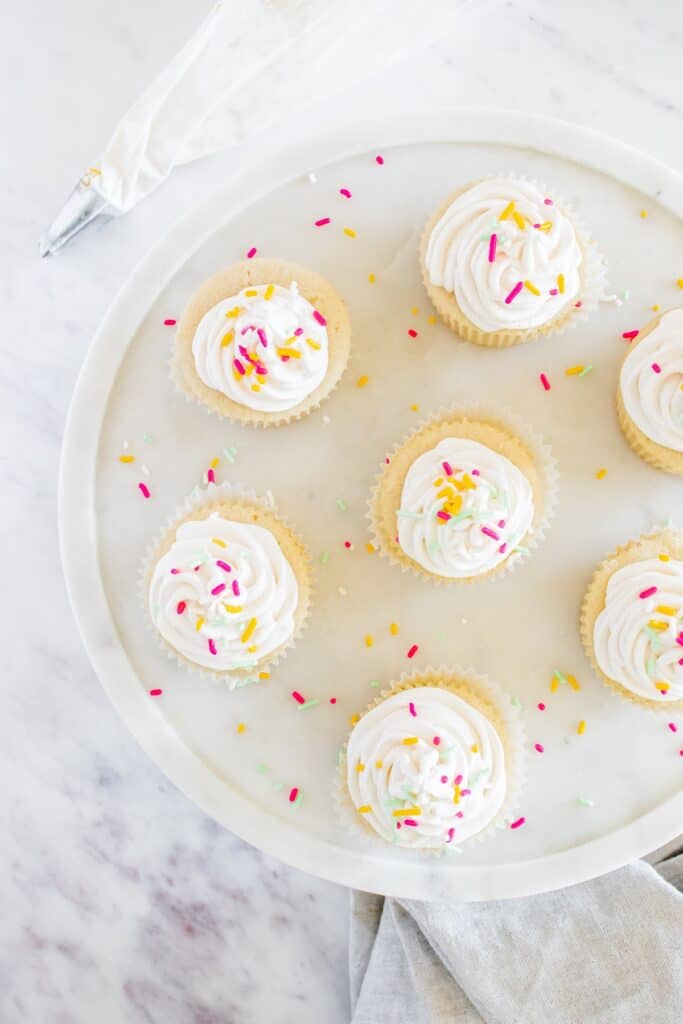 A platter full of cupcakes with dairy free vanilla frosting and sprinkles, next to a piping bag.