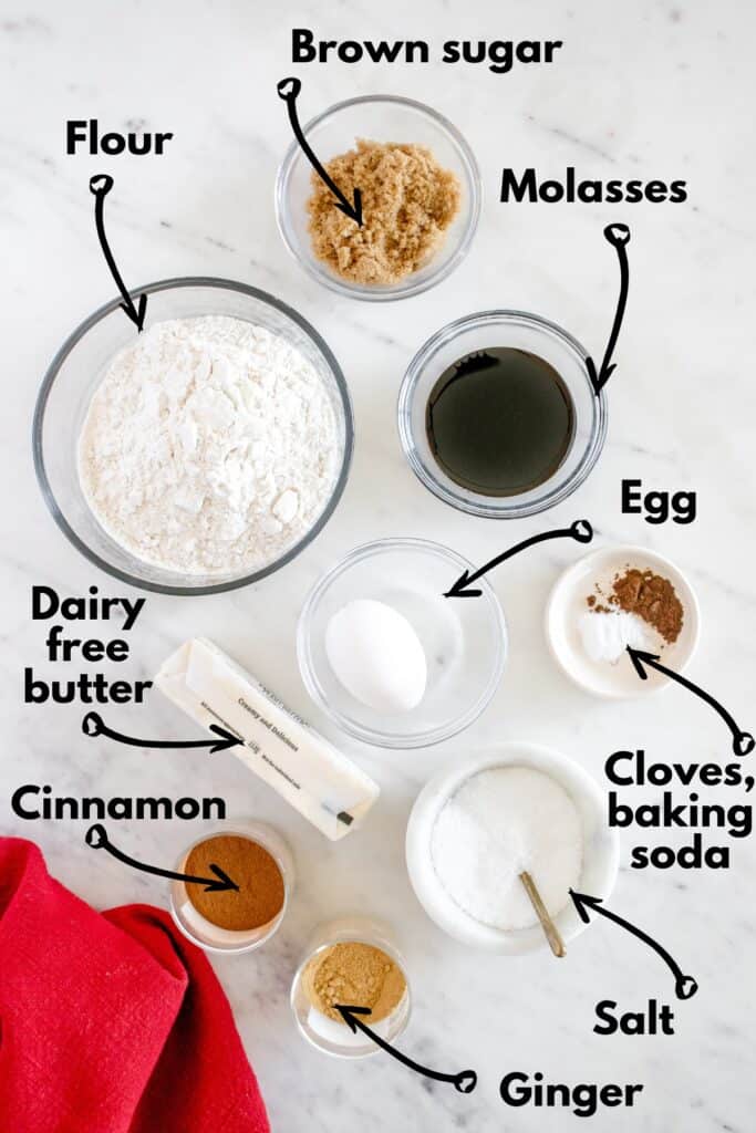 Flour, brown sugar, molasses, dairy free butter, egg, cloves, ginger, cinnamon, and baking soda on a marble countertop.