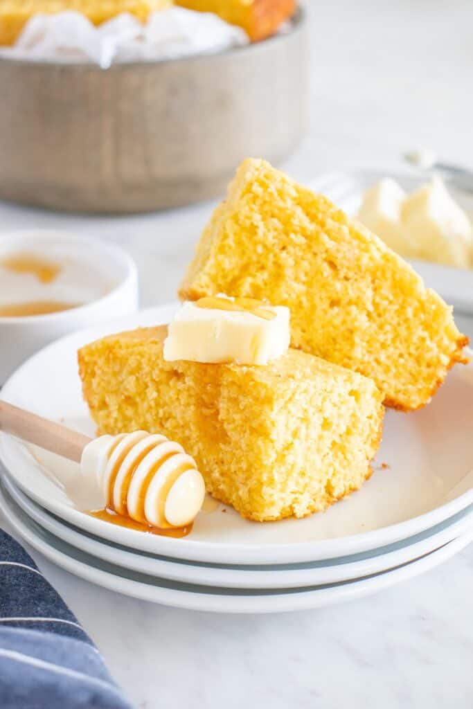 Two pieces of dairy free cornbread on a plate topped with dairy free butter and honey.