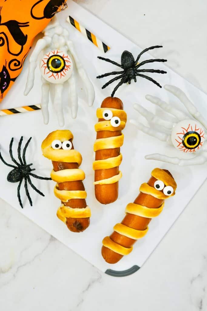 Several mummy hot dogs next to fake spiders, fake eyeballs, and an orange napkin.