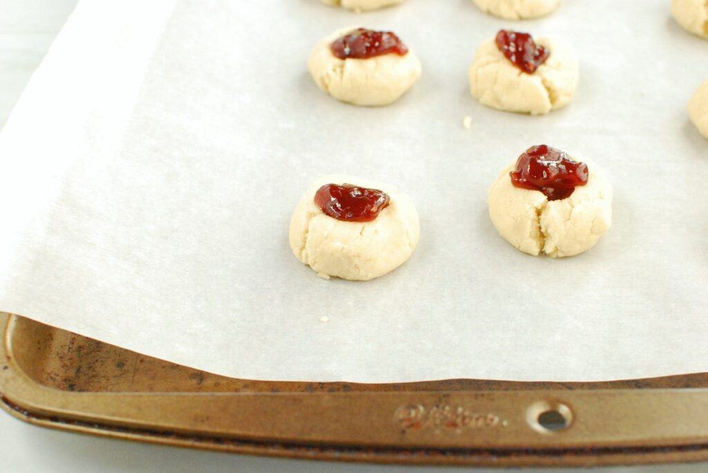 Cookie dough balls on a baking sheet with jam added into the thumbprint spot.