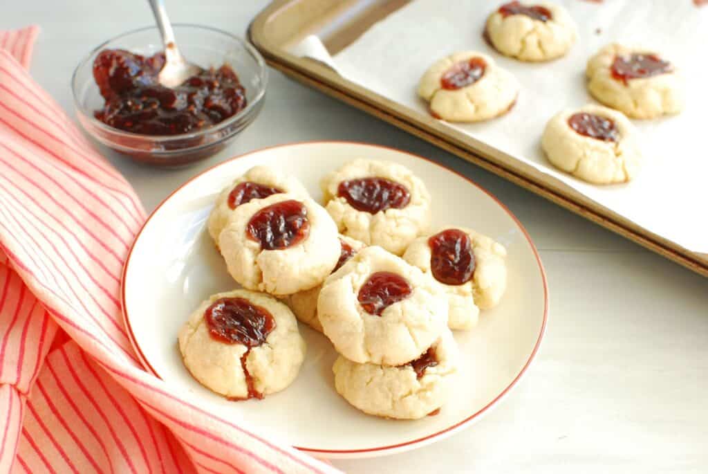 A plate of dairy free thumbprint cookies next to a napkin, baking sheet, and jam.