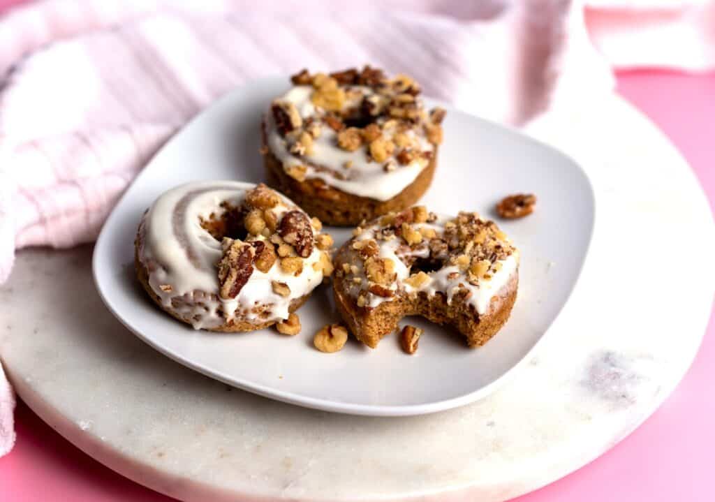 Three dairy free carrot cake donuts topped with frosting and nuts, and one has a bite taken out of it.