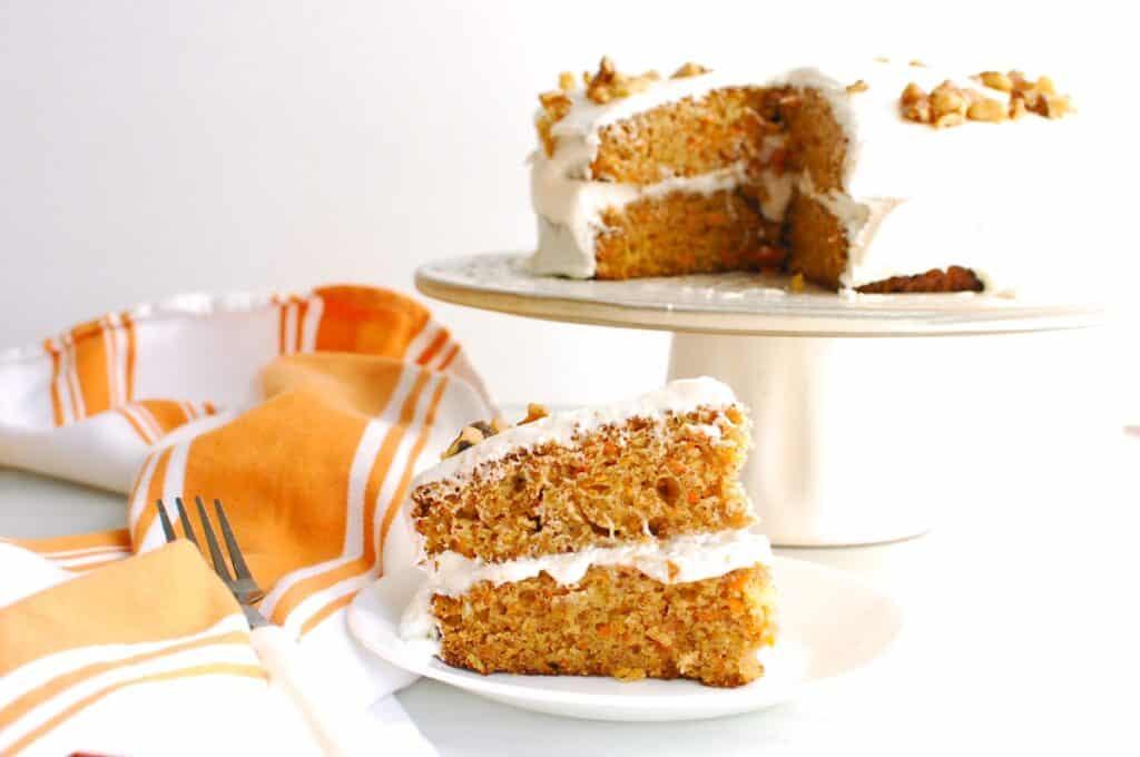 A slice of dairy free carrot cake on a plate with the rest of the cake in the background.