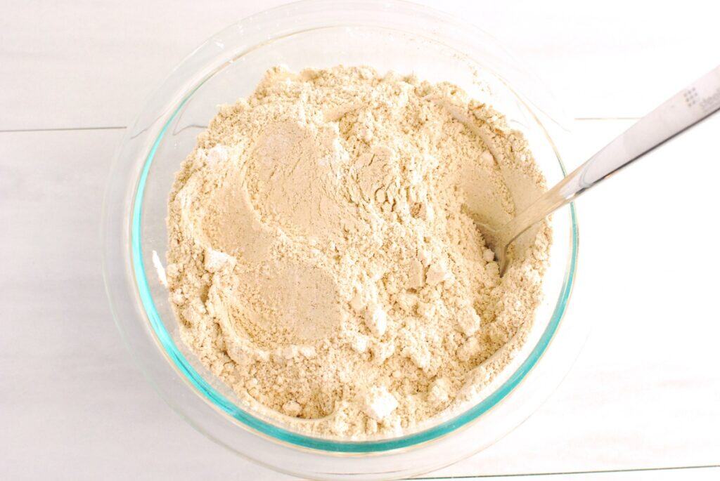 Protein powder, almond flour, oat flour, cinnamon, baking powder, baking soda, and salt mixed together in a glass bowl.