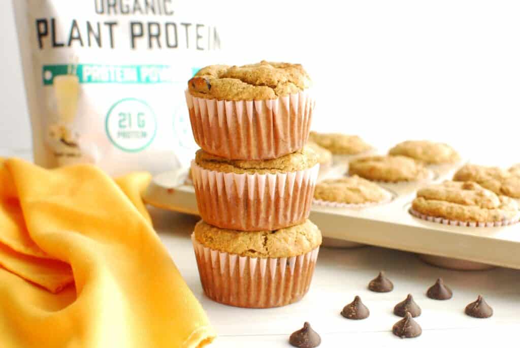 Three dairy free gluten free banana protein muffins stacked on top of each other, with a bag of protein powder in the background.