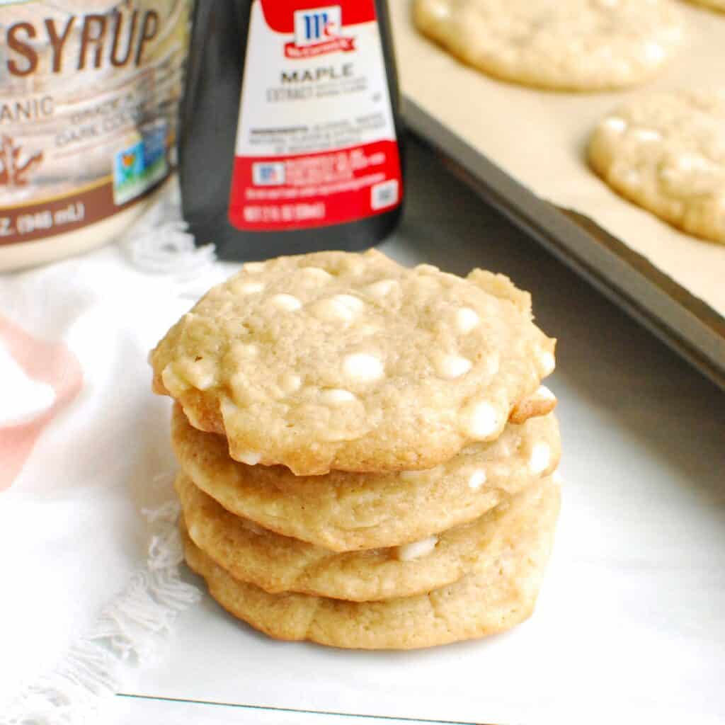 A stack of four dairy free maple white chocolate chip cookies, next to some maple extract and maple syrup.