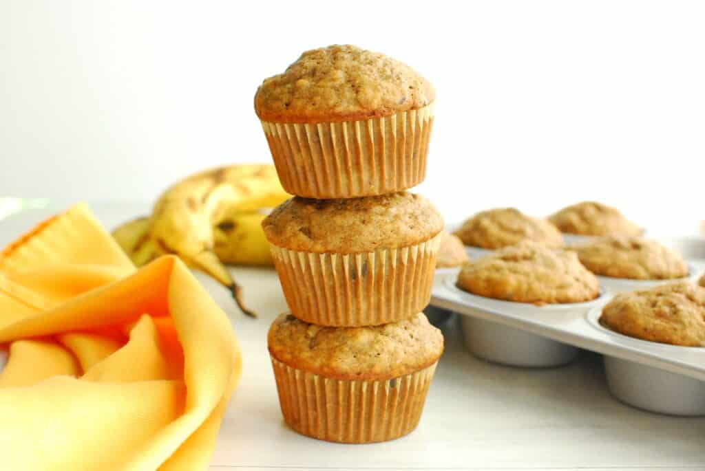 Three chai spiced banana muffins stacked, with additional muffins and bananas in the background.