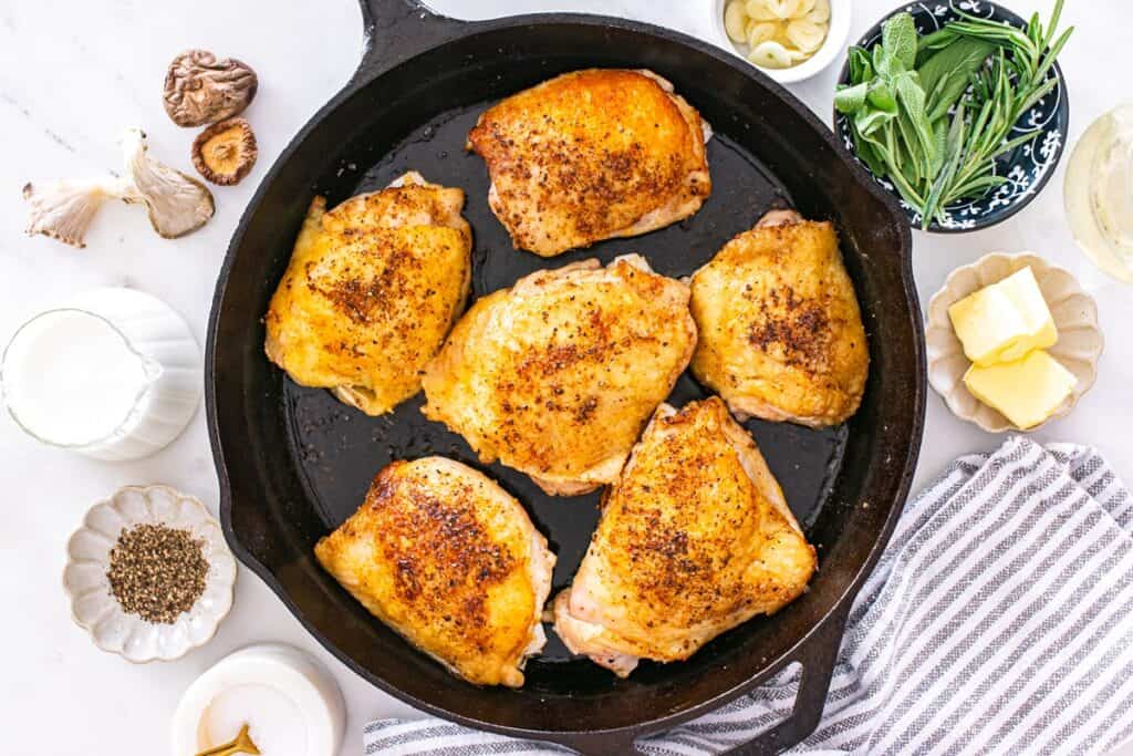 Six chicken thighs cooking in a cast iron skillet.