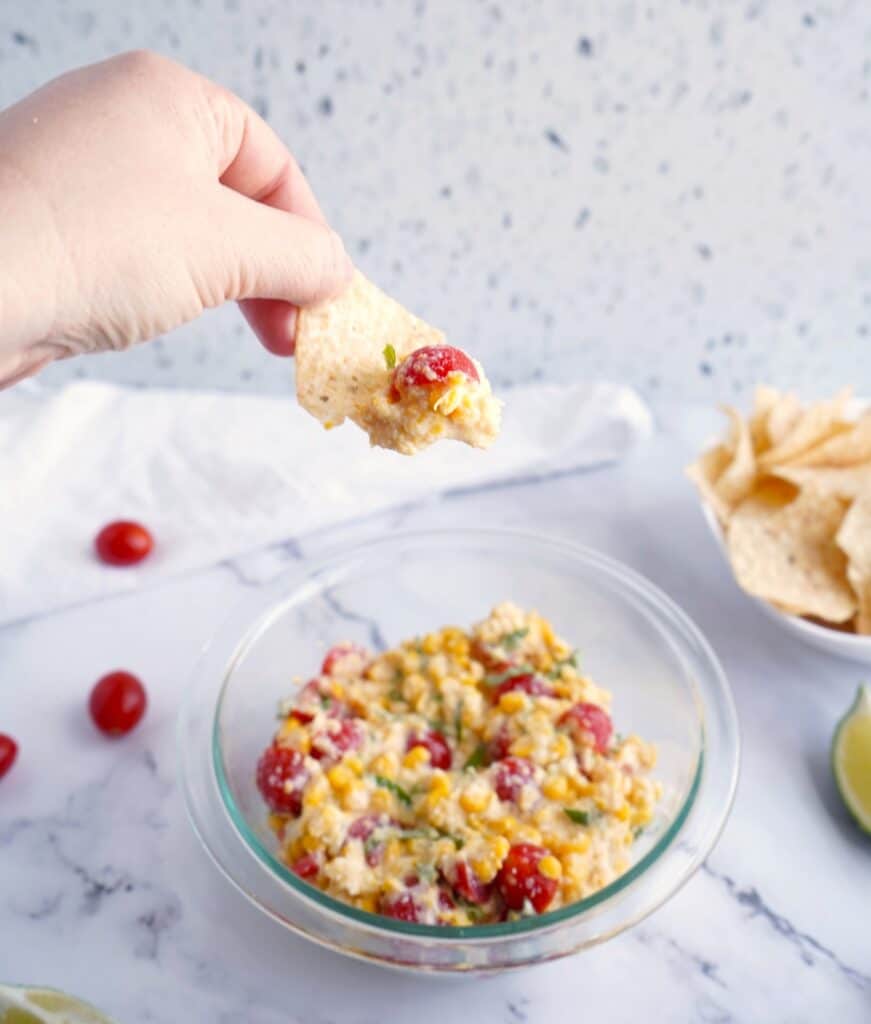 A hand holding a tortilla chip scooping up dairy free corn dip.