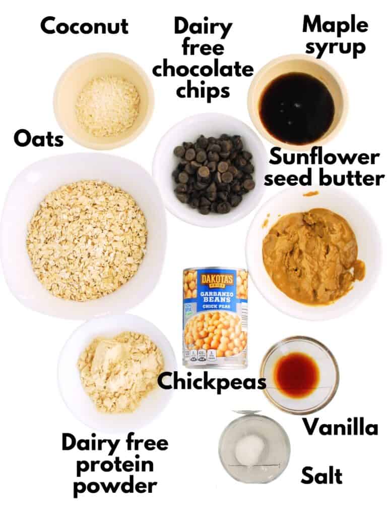 Coconut, chocolate chips, maple syrup, sunflower seed butter, oats, chickpeas, salt, vanilla, and protein powder, all on a white backdrop.