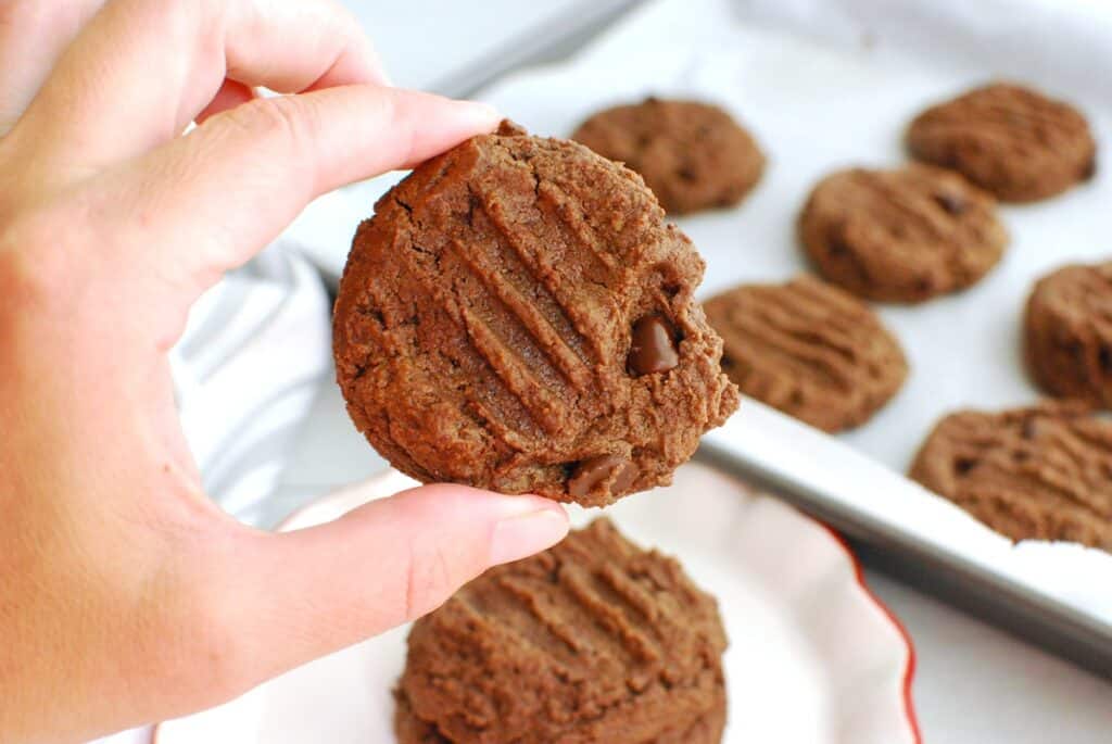 A hand holding a cookie with a baking sheet of cookies in the background.