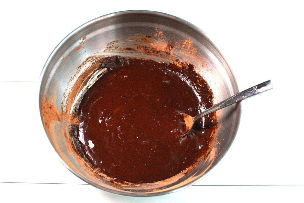 Sugar, cocoa powder, oil, eggs, and vanilla mixed together in a silver mixing bowl.